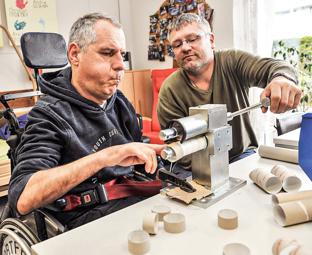 Betreuer hilft Mann mit Behinderung an Kartonmaschine