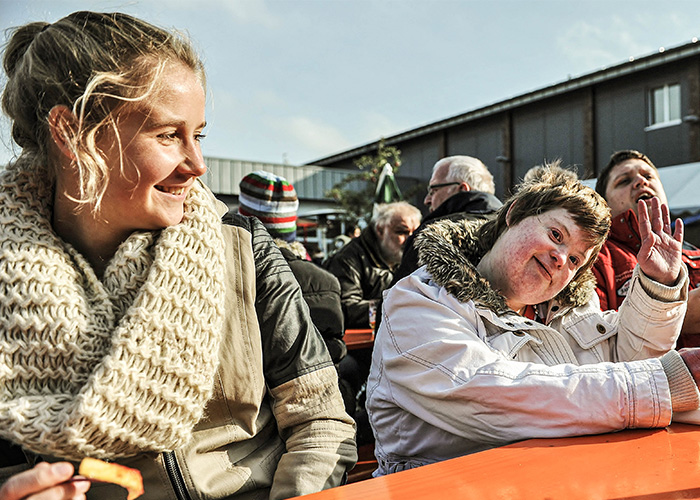 junge Frau sitzt neben Perosn mit Behinderung die in Kamera winkt