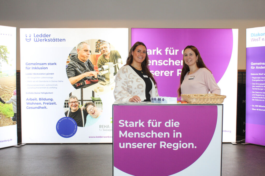 Zwei Frauen stehen an einem Messestand auf einer Ausbildungsmesse. Sie informieren über berufliche Möglichkeiten und tauschen sich mit Ausstellenden und Besuchern aus.